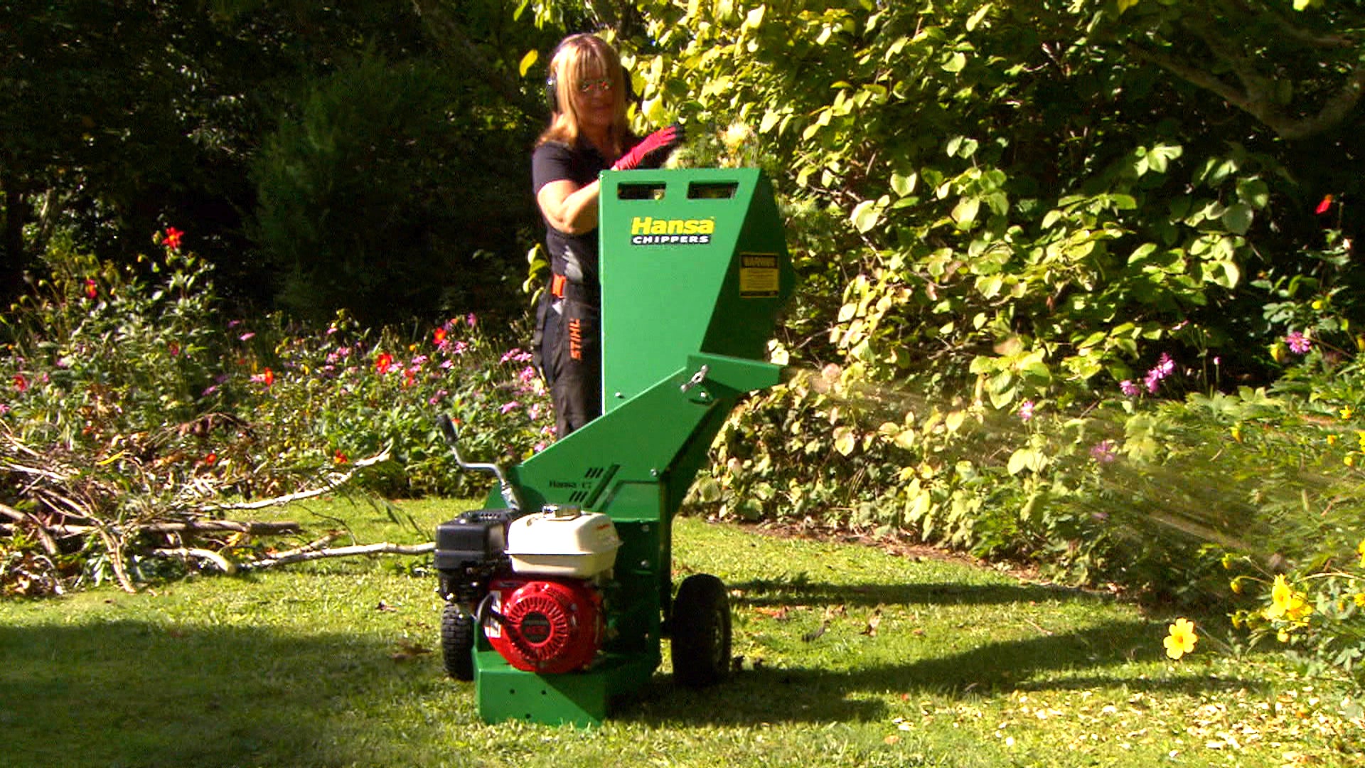 Person in a garden putting branches into a green hansa petrol chipper with a garden in the back ground and flowers. 