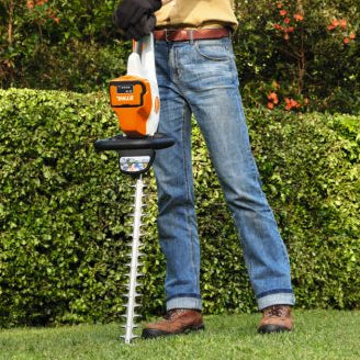Person using a Stihl battery hedge trimmer in a garden setting with greenery and a house in the background.