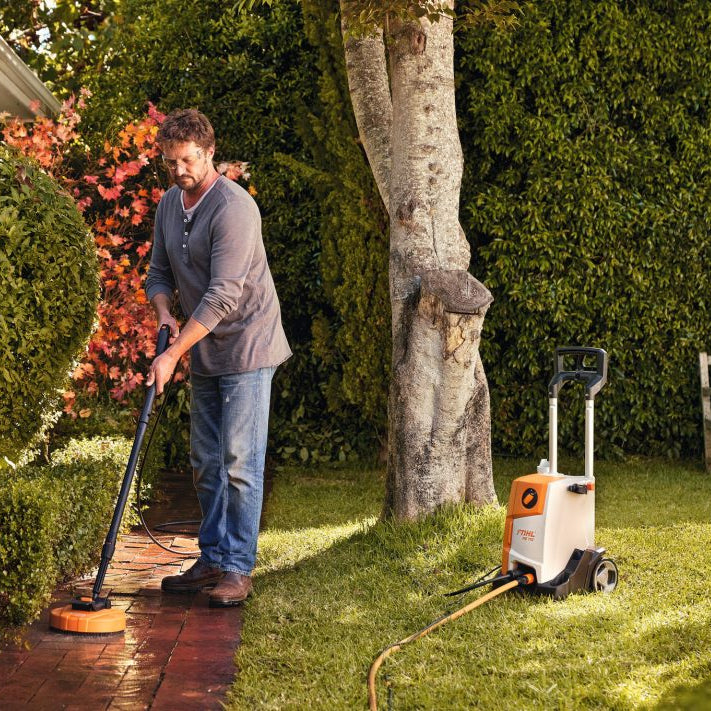 Man using a Stihl Electric power washer on a driveway with garden and bench in the background