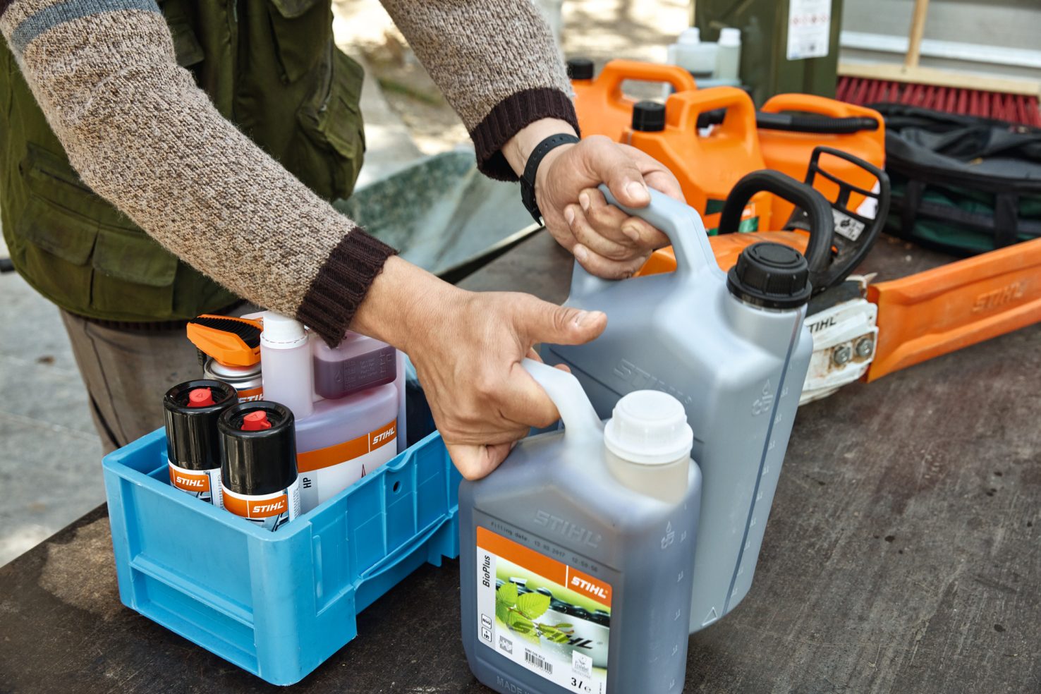 Person holding two bottles of Stihl oil in silver bottles with other stihl accessories and a chainsaw on a table. 